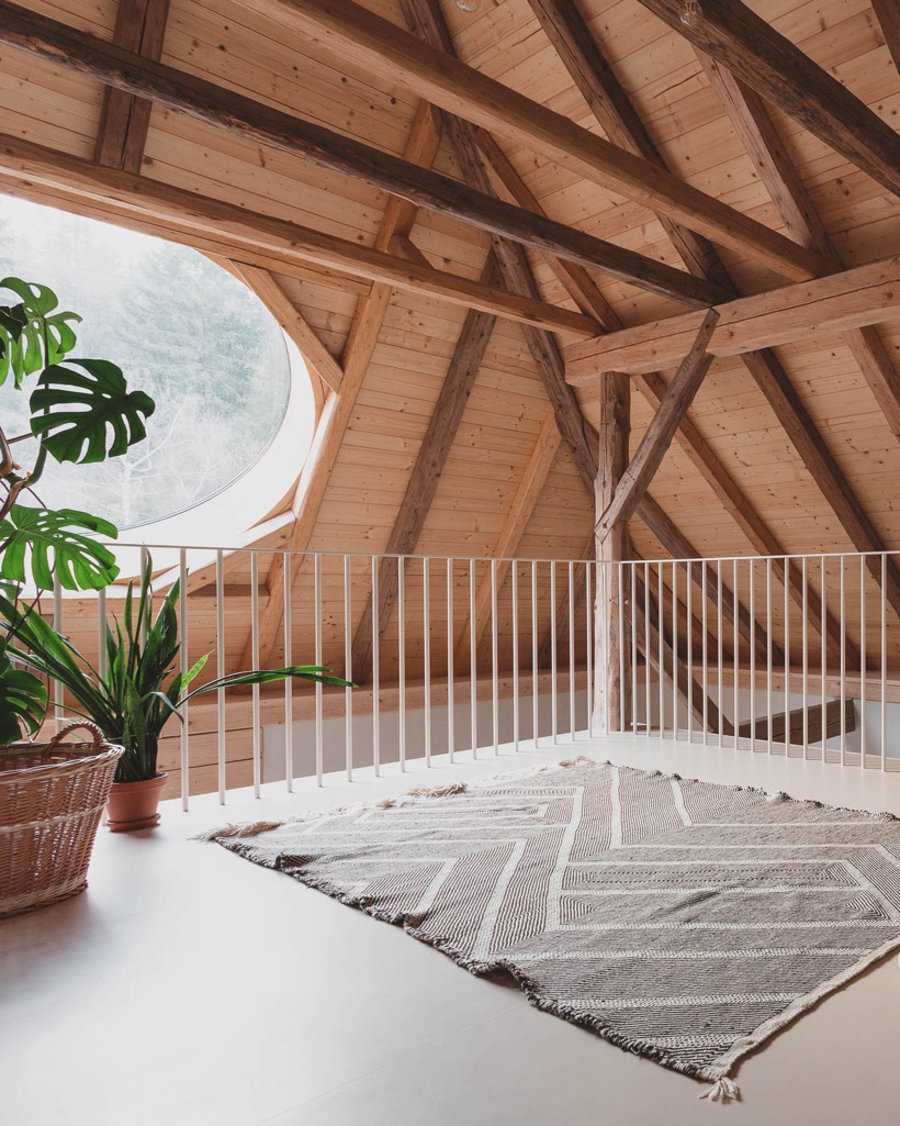 Kruhové okno do lesa, bílé kovové zábradlí galerie, vlněný koberec a monstera v proutěném koši